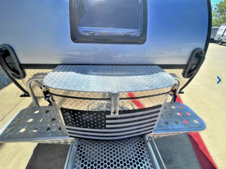 Rear view of a tear-drop RV rental featuring a diamond plate storage box with an American flag design and perforated steps.. T@G XL Boondocks 2023