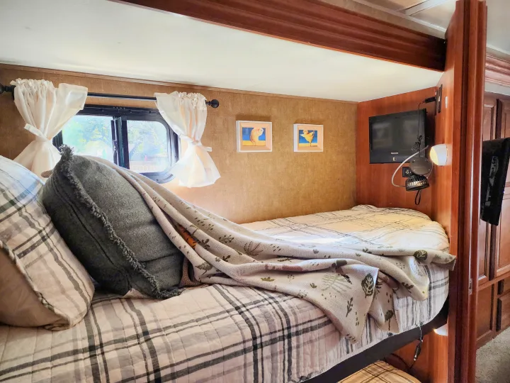 An RV rental's comfy bunk bed setup: plaid bedding, cozy blanket, bright window, charming wall art & TV.. Coachmen Leprechaun 320BH 2014