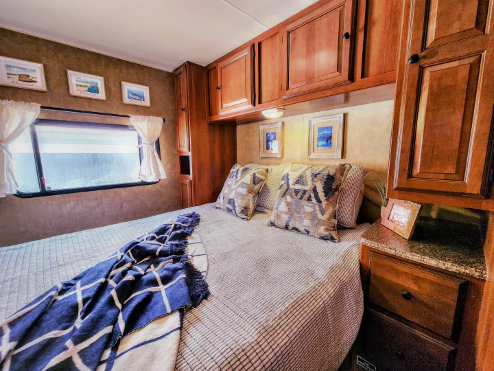 Inviting RV rental bedroom with patterned bed, blue blanket, overhead cabinets, and a window for comfortable stays.. Coachmen Leprechaun 320BH 2014