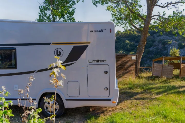 Side view of a white **rv rental** (S340 UP Limited) at a green campsite with trees & wooden facilities.. Benimar Sport 340 UP 2024