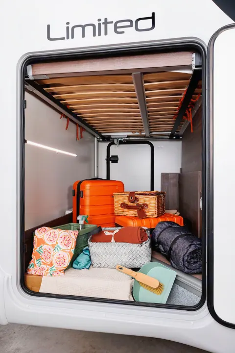 Inside a clean RV rental, the spacious rear garage holds orange luggage, a picnic basket, blankets, and cleaning supplies below the bed.. Benimar Sport 340 UP 2024