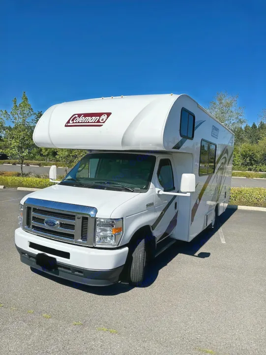Front-angle view of a white Class C Coleman RV rental, a Ford motorhome, parked on asphalt under a clear blue sky.. Coleman 22EQ 2022