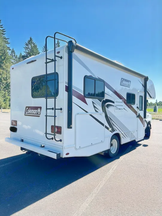 This white Coleman RV rental is ready for adventure. A spacious motorhome with side graphics and a rear ladder, parked on asphalt.. Coleman 22EQ 2022