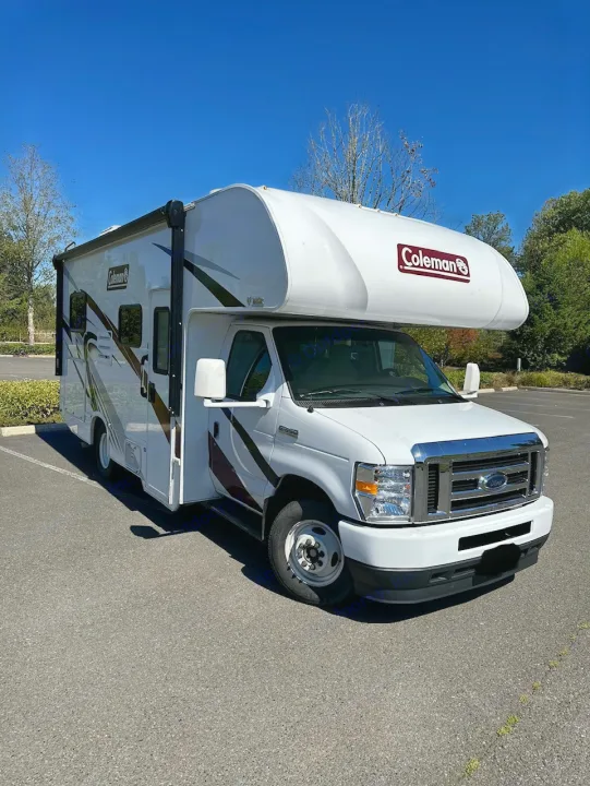 A white Coleman Class C RV rental is parked on an asphalt lot under a clear blue sky, ready for your next road trip.. Coleman 22EQ 2022
