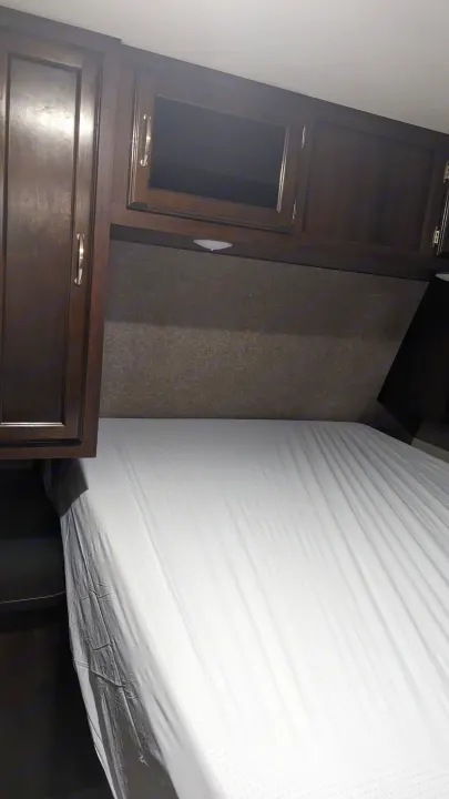 Interior of a clean RV rental bedroom, featuring dark wood cabinets, a grey headboard, and a white mattress.. Jayco Jay Flight 2018