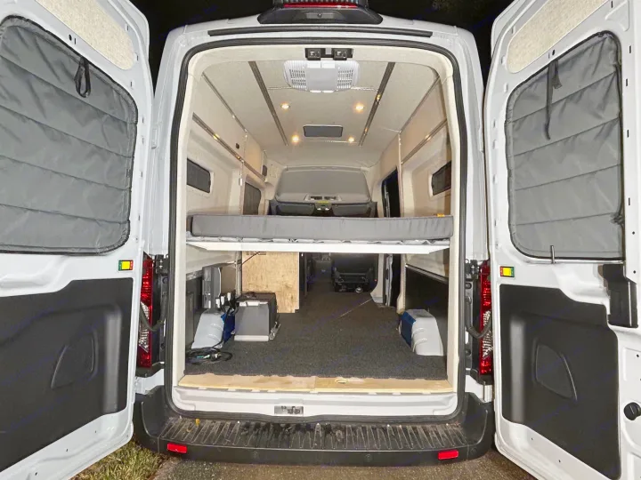 Rear view into a well-lit RV rental camper van interior. Features a comfy bed platform, lower cargo area, and insulated doors.. Ford Transit 2023