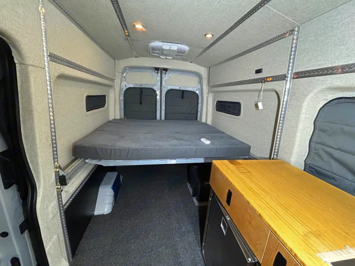 RV rental: Van interior with a raised bed, under-bed storage, and walls lined with light-colored fabric and mounting tracks.. Ford Transit 2023