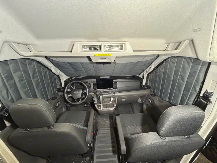 Inside an RV rental van: a clean front cabin with dashboard, steering wheel, two bucket seats, and quilted window coverings.. Ford Transit 2023