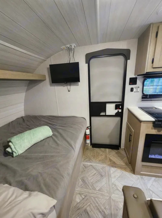 Compact RV rental interior showcasing a modern bed, wall-mounted TV, small kitchen, and patterned floor. Ideal for travel.. Other Other 2024