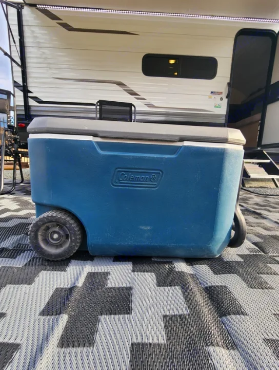 A wheeled teal Coleman cooler sits on a patterned mat in front of an RV rental, which has an awning with LED lights.. Other Other 2024