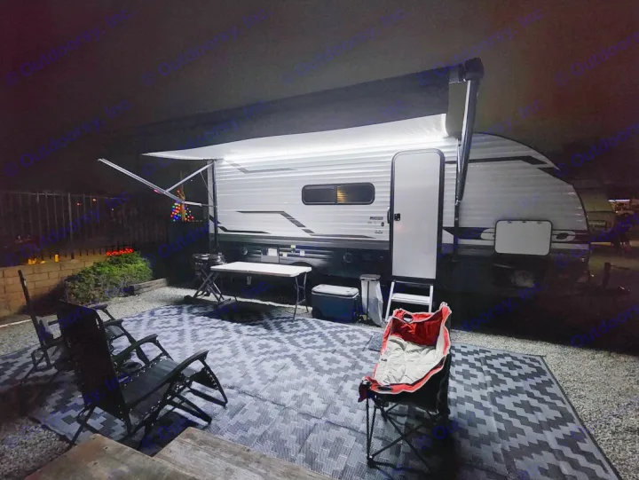 Cozy night at an RV rental campsite with a lit awning, outdoor mat, and chairs for relaxation.. Other Other 2024