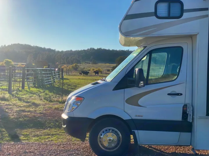 White RV rental on gravel, parked in a sunny rural landscape with cows, fences, and forested hills.. Forest River Coachmen Prism 2014