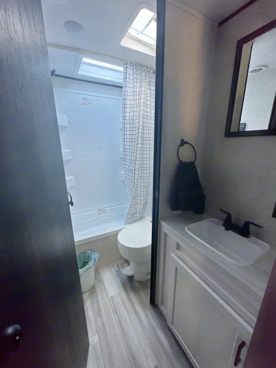 RV rental bathroom featuring a white shower/tub combo, toilet, vanity with sink, and mirror. Gray wood-look flooring adds style.. Other Other 2024