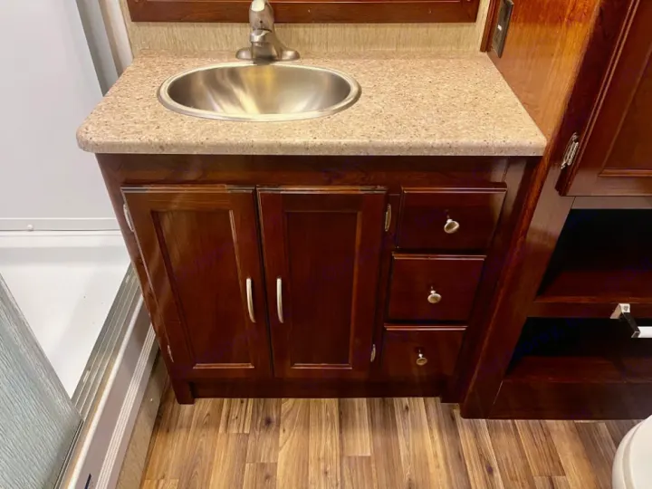 RV rental's bathroom vanity: wood cabinets, granite-look countertop, stainless sink, and faucet. Adjacent shower and wood-look floor.. Coachmen Mirada 2018