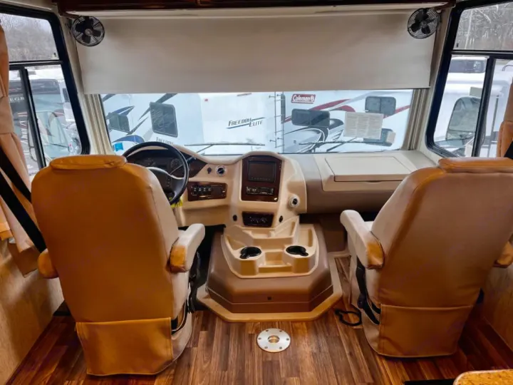 Spacious RV rental cockpit with tan captain's chairs, steering wheel, and dashboard. View through windshield shows other RVs.. Coachmen Mirada 2018