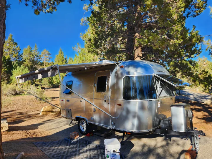 A sleek Airstream travel trailer, set up at a wooded campsite with awning & lights, perfect for an rv rental getaway.. Airstream Bambi 2023