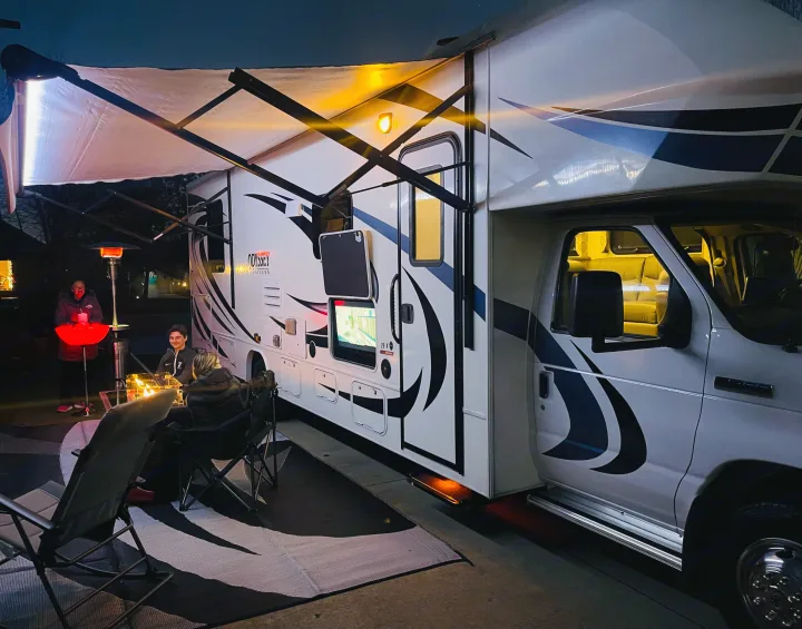 Night scene with an RV rental, people enjoying an outdoor living setup with an awning, heater, and TV.. Entegra Coach Odyssey 2020