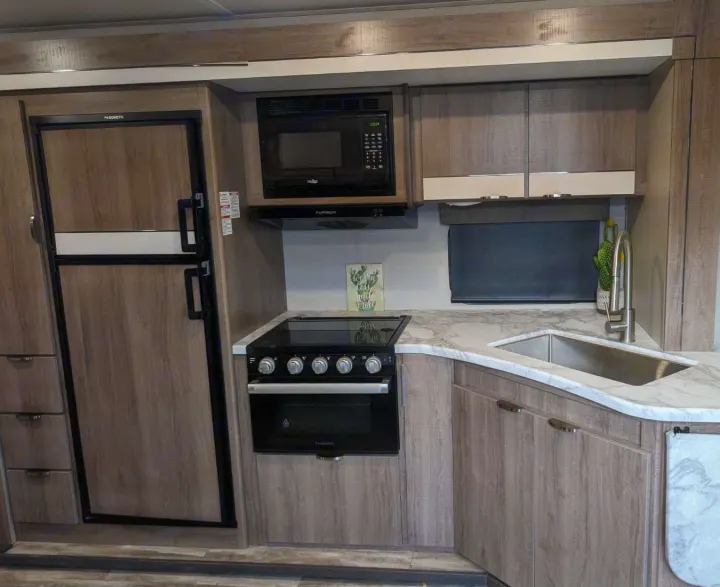 Modern RV rental kitchen featuring a Dometic fridge, Furrion microwave and stove, marble-look counters, and wood-grain cabinets.. Grand Design Imagine XLS 2021