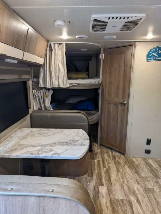 Inside this RV rental: view of a dinette with marble-look table, bunk beds with curtains, AC, and wood-patterned floor.. Grand Design Imagine XLS 2021