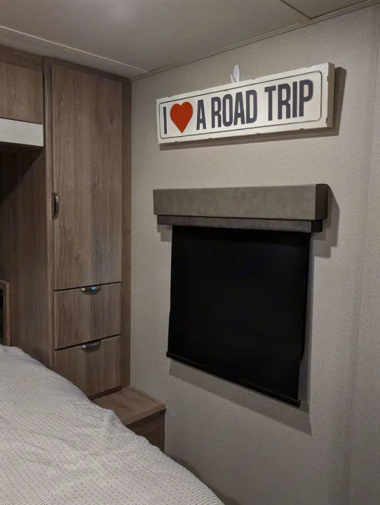 RV rental bedroom interior: wooden cabinets, bed, and "I ❤️ A ROAD TRIP" sign above a window with a black shade.. Grand Design Imagine XLS 2021