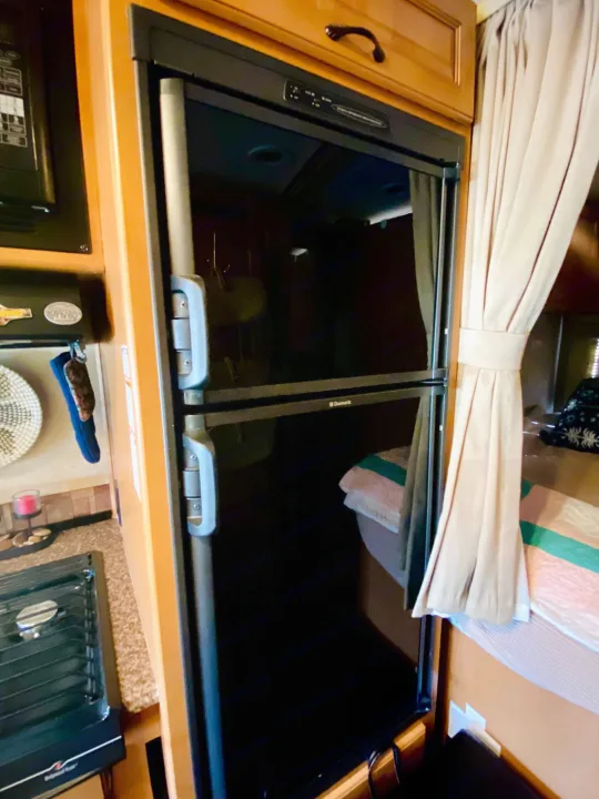 Inside an rv rental, a large Dometic refrigerator/freezer is built into wooden cabinetry, next to a kitchenette and bed.. Forest River Coachmen Prism 2014