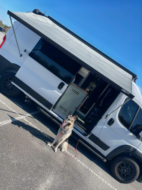 Happy dog on leash beside a white RV rental camper van with an extended awning, open side door, ready for adventure.. 27North 27North Class B 2023