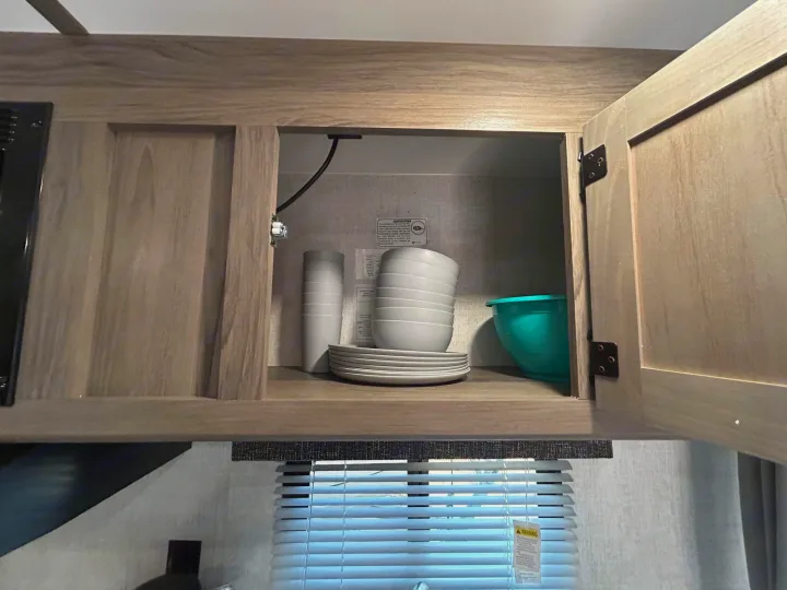 RV rental kitchen cabinet open with neatly stacked gray dishes, cups, and a teal bowl, ready for your travels.. Gulf Stream Ameri-Lite 2022