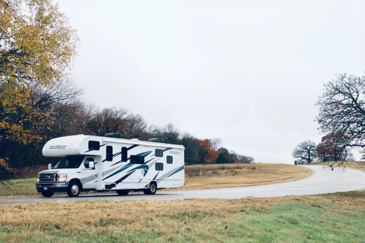 White "Freedom Elite" RV rental on a winding road with autumn trees and grass under a cloudy sky, suggesting a scenic road trip.. Thor Freedom Elite 2023