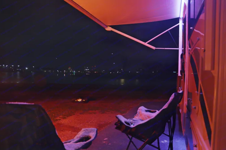 Night view from an RV rental: illuminated awning, campfire, and distant city lights over the water. Perfect for camping!. Thor Freedom Elite 2023