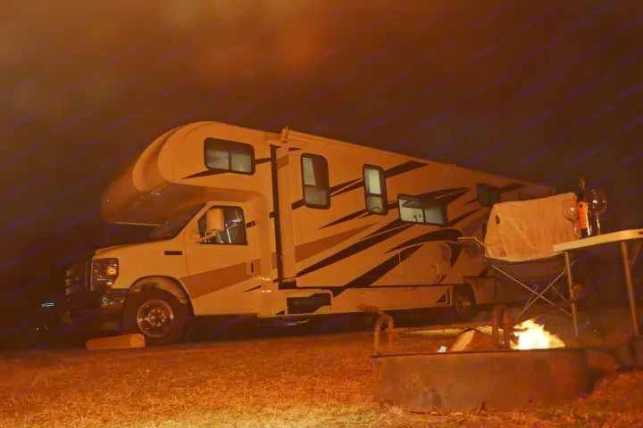 Cozy night scene: a white RV rental parked under a dark sky, illuminated by a glowing campfire in the foreground.. Thor Freedom Elite 2023