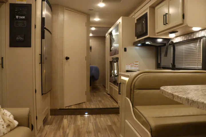 Interior view of an RV rental with a beige dinette, kitchen area, refrigerator, and hallway leading to bunk beds.. Thor Freedom Elite 2023