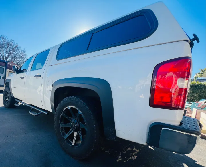 White pickup truck with camper shell, parked by an RV. Offers versatility for your next travel adventure or RV rental trip.. Chevrolet Silverado 1500 2009