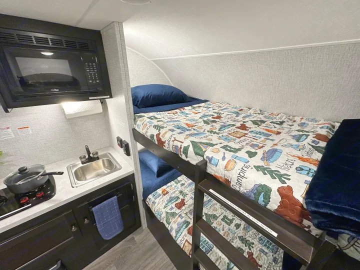 RV rental interior: a compact kitchen area with microwave, sink, and stovetop, adjacent to cozy bunk beds with themed quilts.. Braxton Creek Bushwhacker Plus 2022