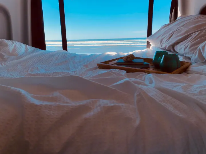 Cozy RV rental bedroom offers stunning ocean views! White bed with a tray, books, and glasses for a serene escape.. Winnebago Revel 2022