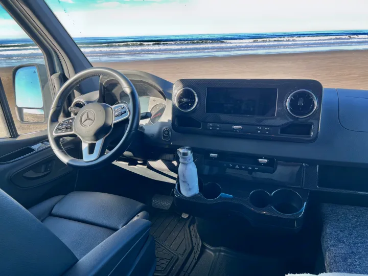 Panoramic beach view from a modern RV rental dashboard, featuring the steering wheel, controls, and infotainment screen.. Winnebago Revel 2022