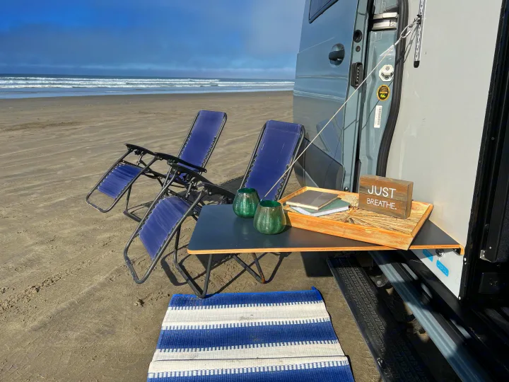 Peaceful RV rental beach day. A gray van with an attached table, two blue chairs, and a mat facing calming ocean waves.. Winnebago Revel 2022
