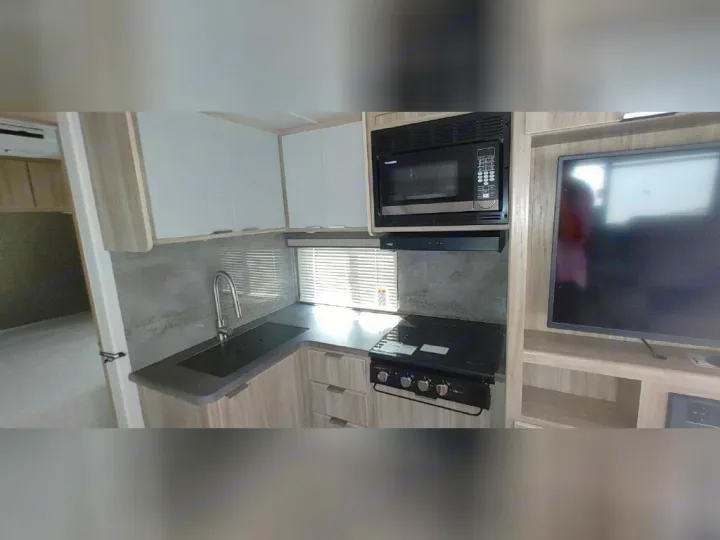 A compact RV rental kitchen features a sink, stovetop, microwave, white cabinets, and a TV setup, ideal for travel.. Winnebago 2301BHS 2022