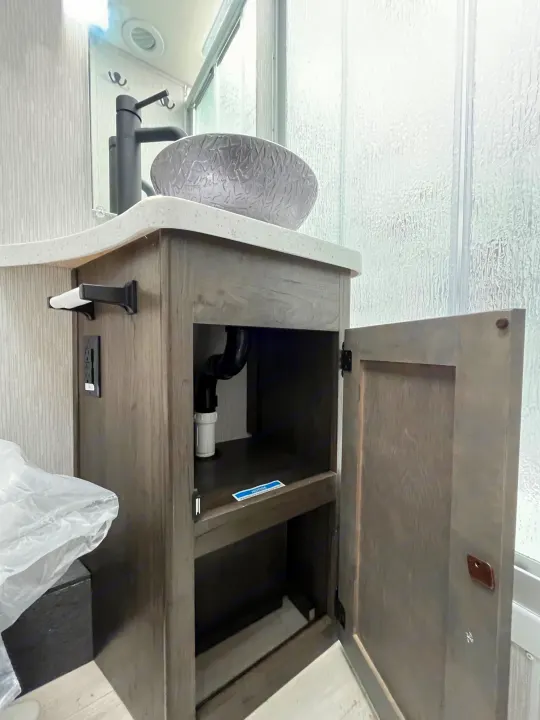 Modern RV rental bathroom vanity with grey vessel sink, black faucet, open cabinet showing plumbing, and shower beside.. Coachmen Prism Elite 2024