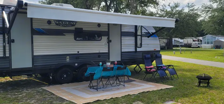 Relaxing RV rental setup: a travel trailer with an extended awning, outdoor rug, table, chairs, and fire pit on green grass.. Forest River Salem Cruise Lite 2021