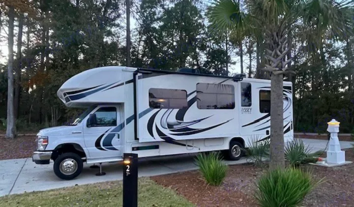 White Class C RV rental with black and grey decals parked at a paved campground site with trees and a utility pedestal.. Entegra Coach Odyssey 2020