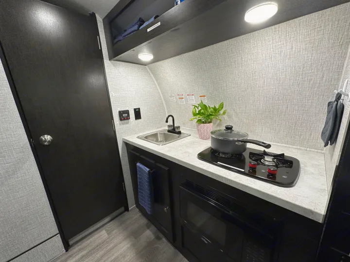 RV rental interior showcasing a compact kitchenette with a sink, two-burner stove, microwave, and overhead storage.. Braxton Creek Bushwhacker Plus 15DS 2022