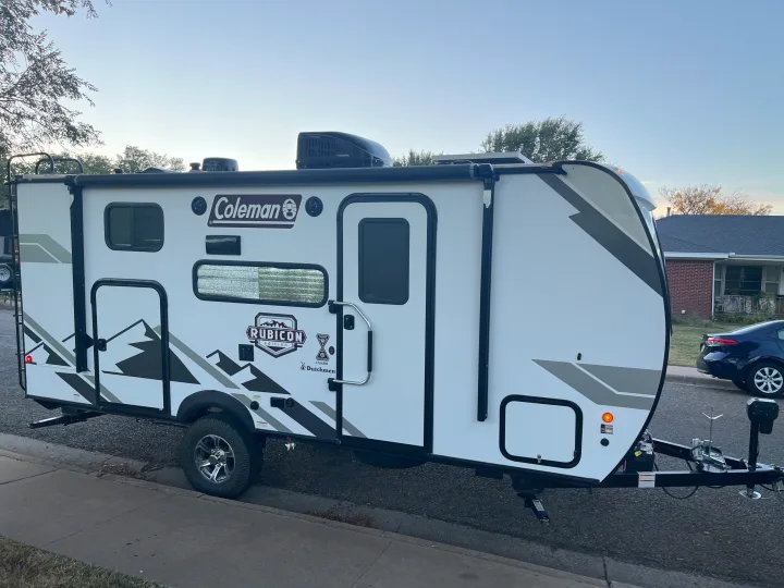 A white Coleman Rubicon travel trailer with mountain graphics is parked, showcasing features like its awning, ideal for RV rental.. Dutchmen Coleman Rubicon 2022