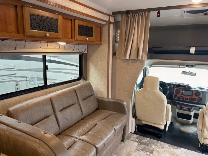 Spacious RV rental interior with a brown sofa, wood cabinets, and the front cab area with an overhead bunk.. COACHMEN LEPRECHAUN 2019