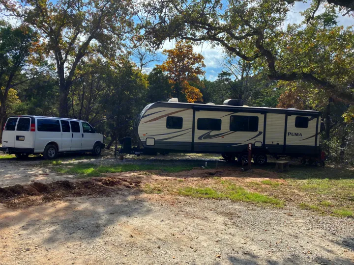 Enjoy nature with this rv rental! A white van and travel trailer are parked on a sunny, wooded campsite.. Forest River Palomino Puma 2018