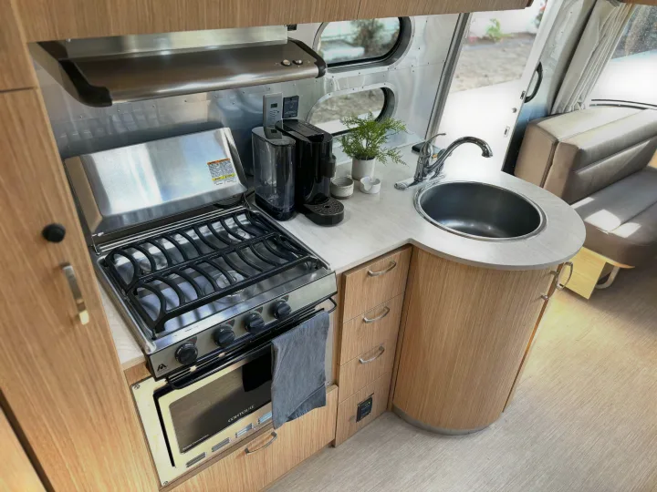 This modern RV rental kitchen features a stainless steel stove, oven, sink, and coffee maker within light wood cabinets.. Airstream Flying Cloud 2017