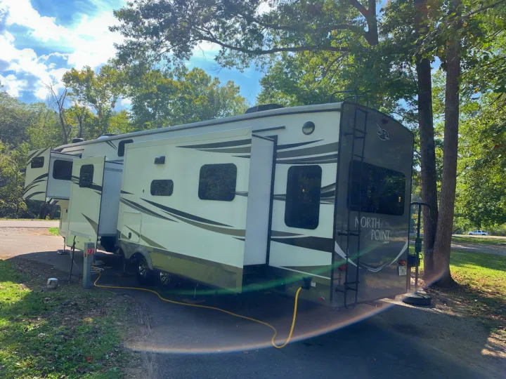 A large white and grey fifth wheel RV rental is parked with slide-outs extended at a sunny, tree-lined campsite. A hose is connected.. Jayco North Point 2021