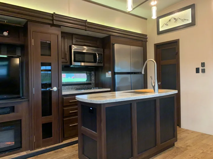 Modern RV rental interior featuring a dark wood kitchen, island, stainless steel appliances, and living area with a TV.. Jayco North Point 2020