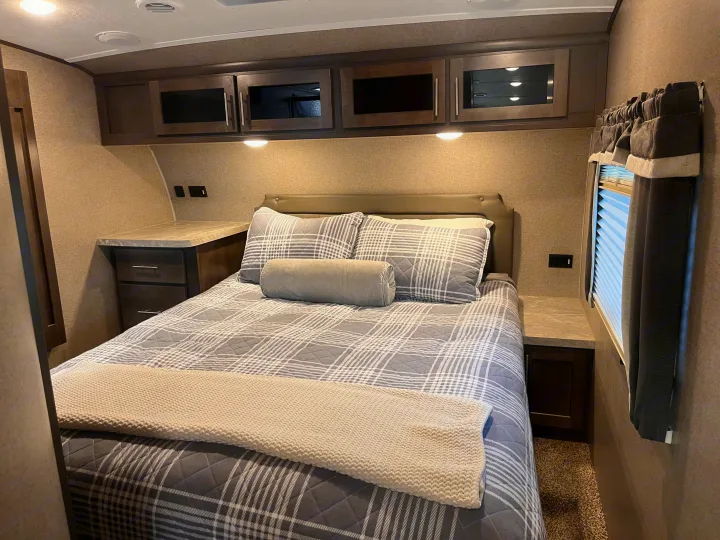 This inviting RV rental bedroom features a made bed with plaid comforter, overhead cabinets, and bedside tables.. Forest River Rockwood Ultra Lite 2020