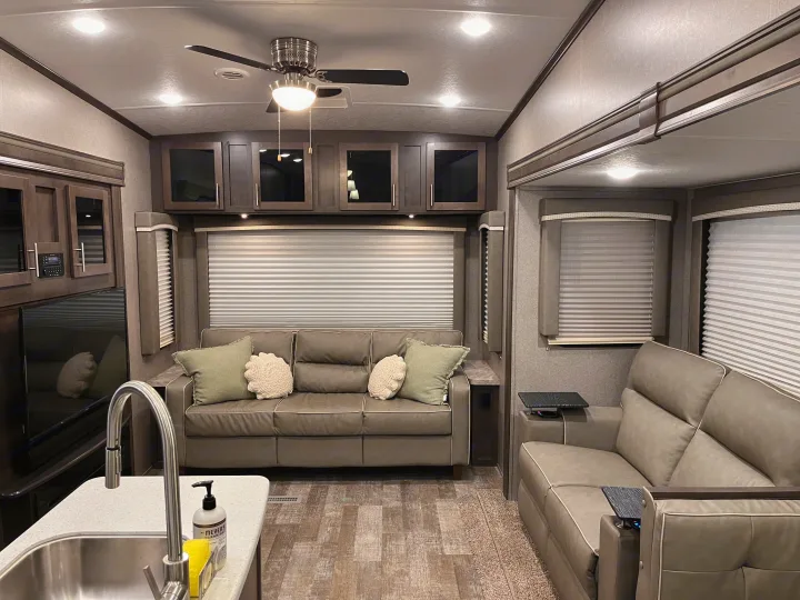 Inside a modern RV rental: A well-lit living area with gray sofas, overhead cabinets, ceiling fan, and a kitchen sink.. Forest River Rockwood Ultra Lite 2020