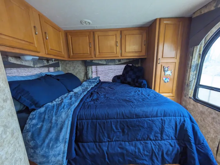 Cozy RV rental bedroom with blue bedding, overhead wood cabinets, and an arched window view.. Coachmen Freelander Deluxe/Premier 2005
